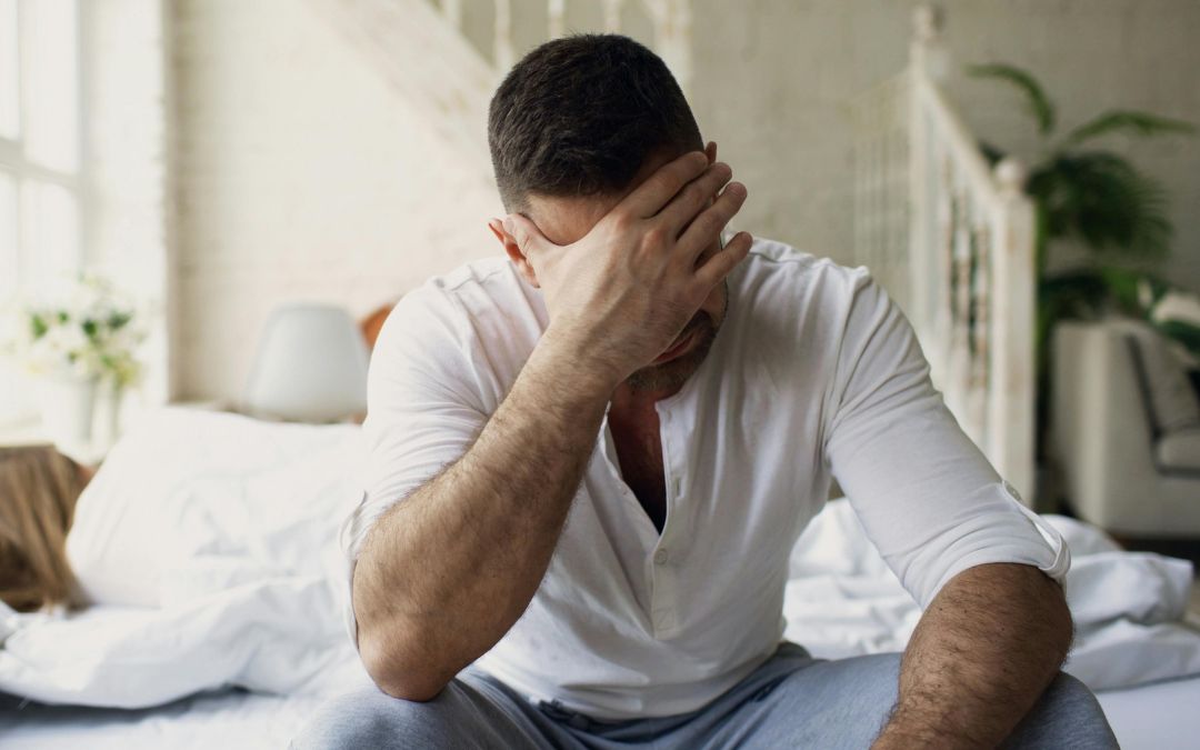 Homme assis sur un lit, gêné par une cage de chasteté inconfortable